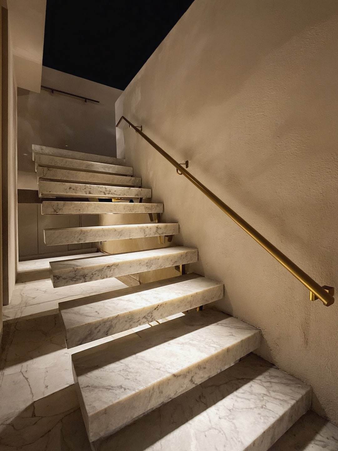 Floating travertine stair treads with a slim bronze handrail against a plaster wall.