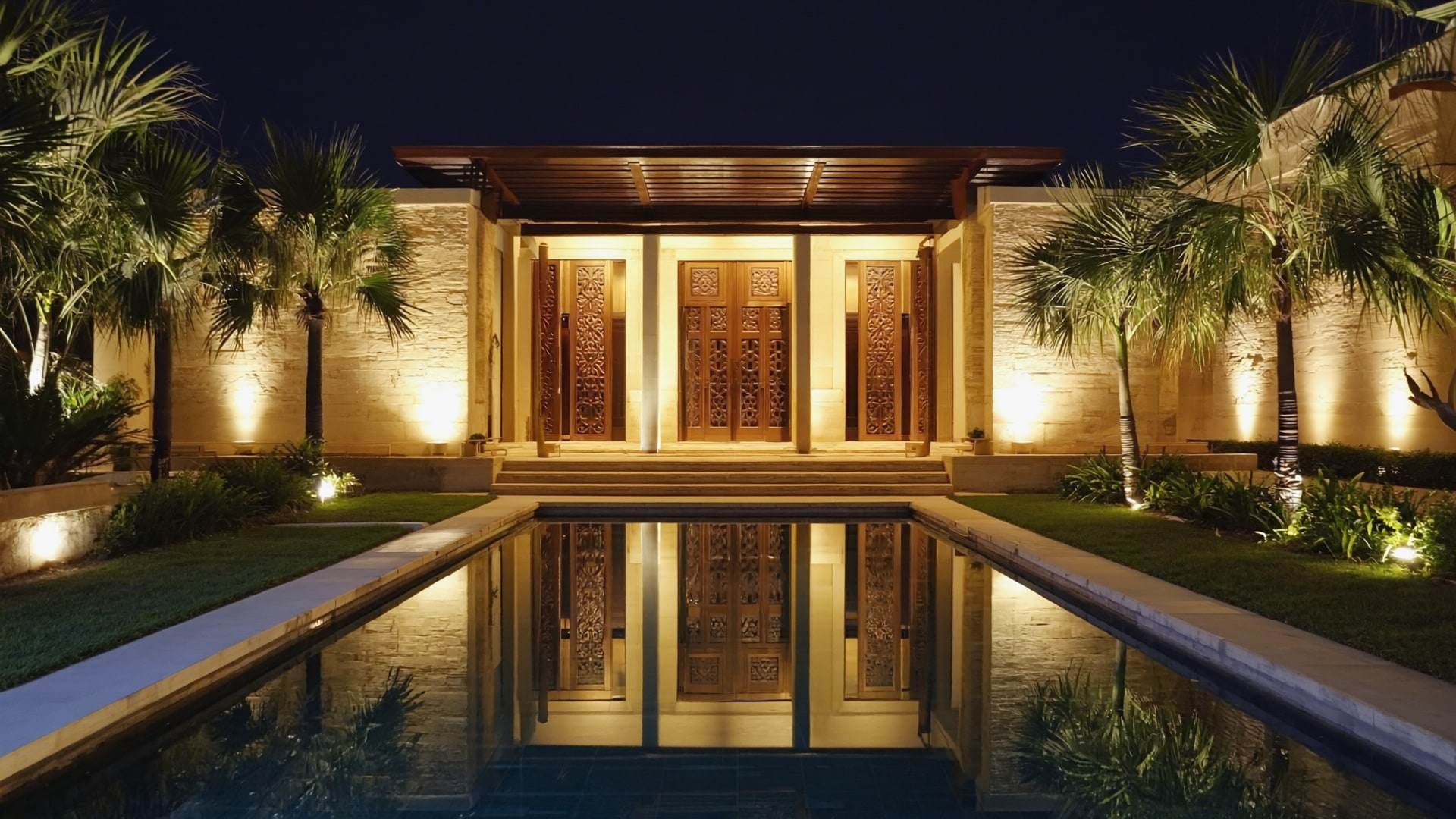 The arrival court of a family estate at night: a long pool leading to carved teak doors.