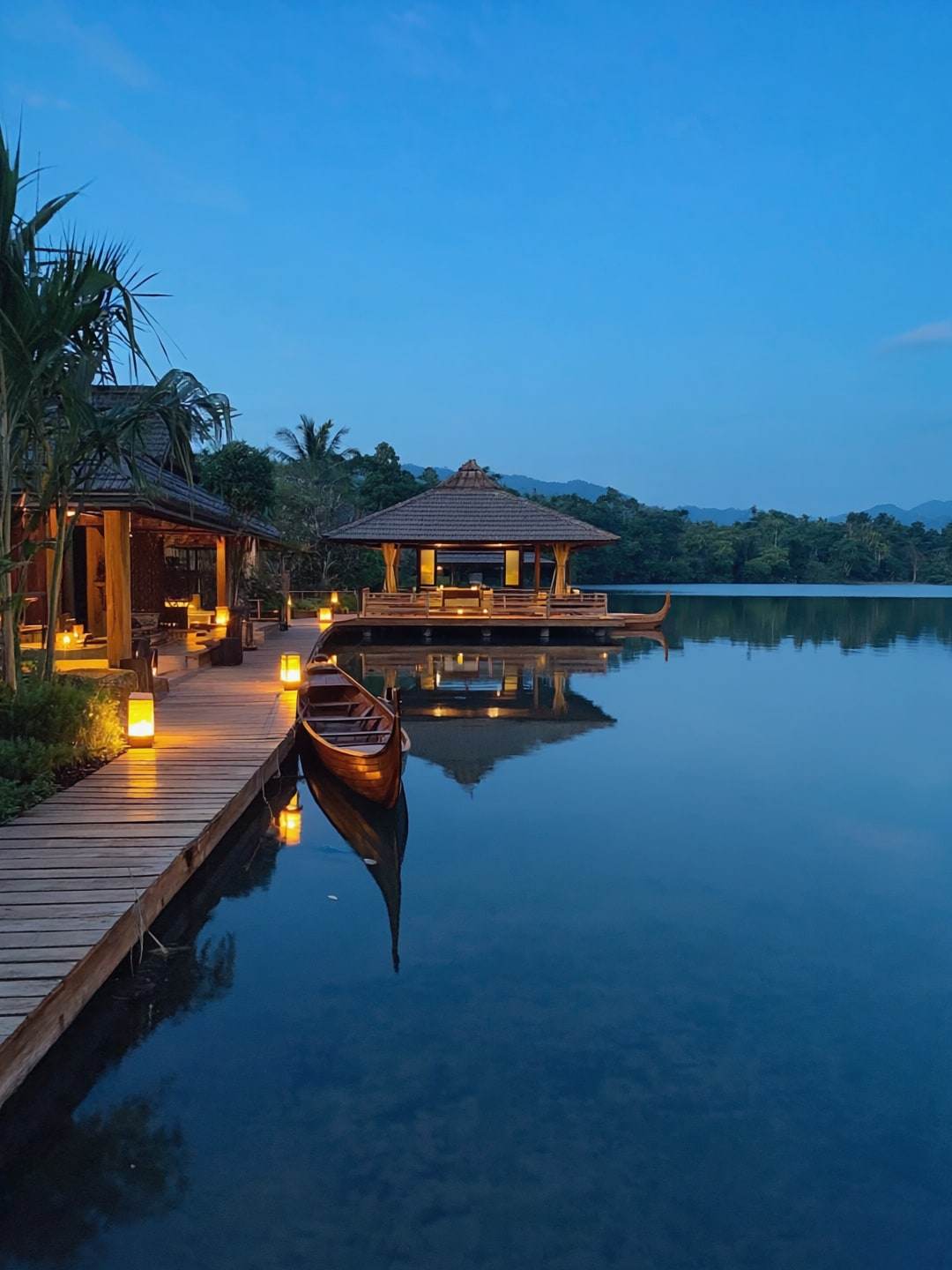 A timber jetty and floating pavilion lit by lanterns at blue hour.
